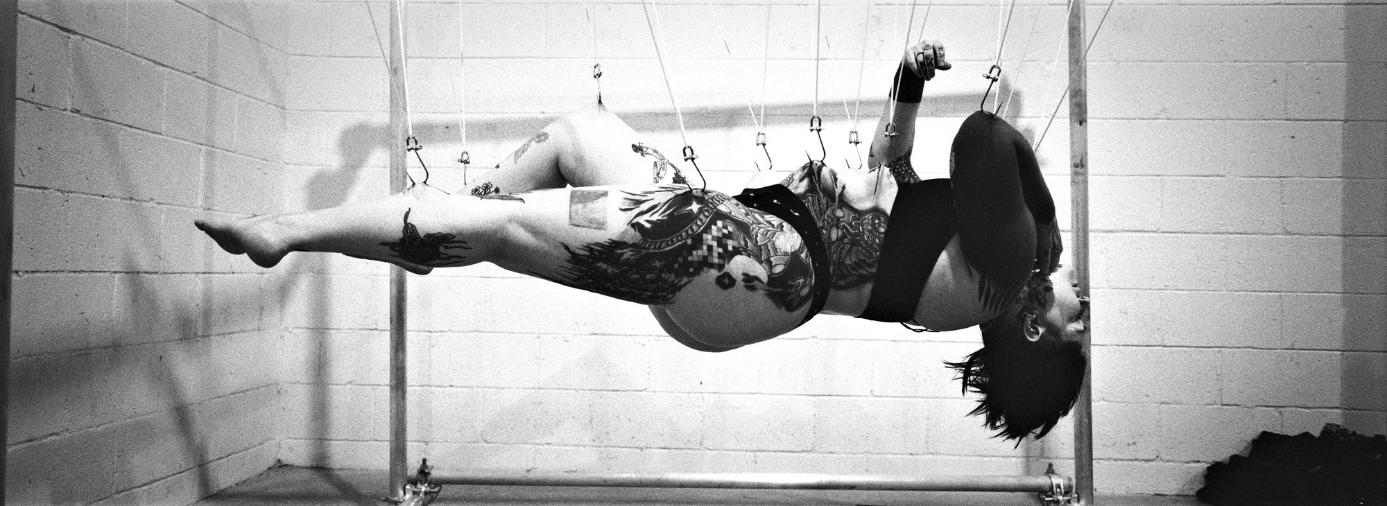 A tattooed person suspends from hooks, laying flat, one leg higher than the other. Their head is back, and they seem to be smiling, dark hair dangling like an anime character.