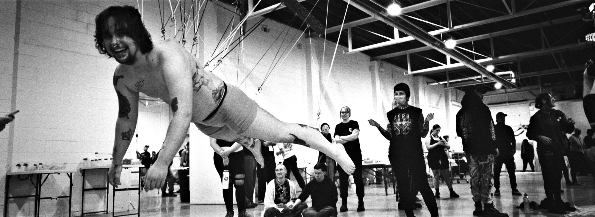 A black-and-white photo of a person mid-air in a Superman-style body suspension pose, supported by multiple hooks in their back and legs, smiling joyfully toward the camera. They are suspended horizontally in a large indoor space with high ceilings and visible rigging. A group of onlookers—some seated, some standing—watch with expressions of admiration, amusement, and support. The atmosphere is lively and communal, capturing a moment of shared experience and transformation.