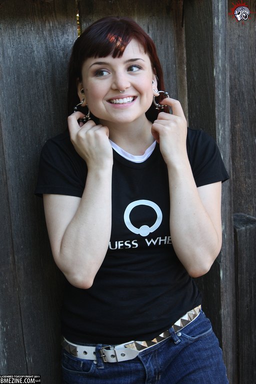 A smiling young woman with red bangs pulls on her hoop earrings and looks off to the side, her t-shirt features a captured bead ring and the words, "Guess Where"