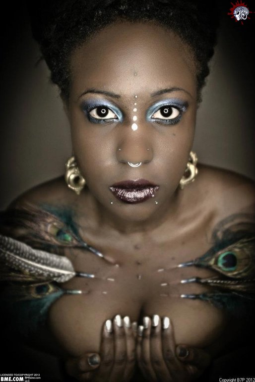 A close portrait of a woman of color with tattoos, and many piercings including play piercings in her chest with peacock feathers attached,  looking directly into the camera.