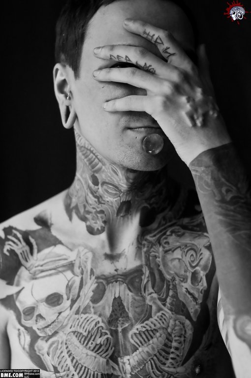 A heavily tattooed, pierced and implanted person covers their face with their hand. Monochrome.