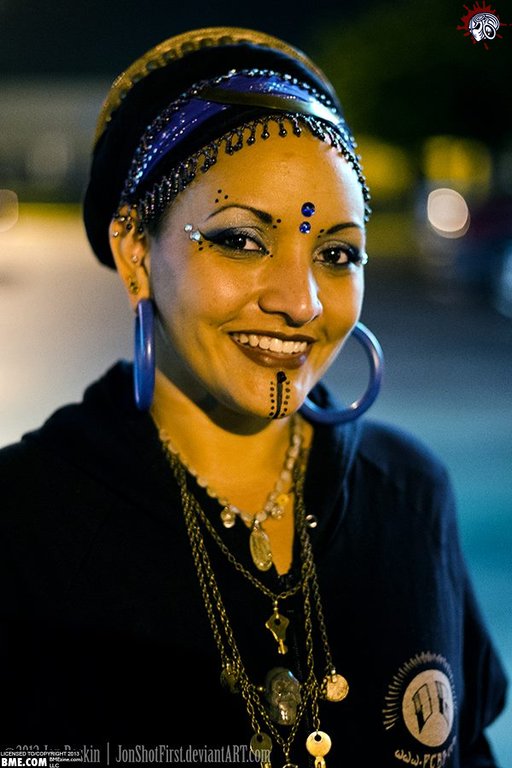 A woman of color smiles into the camera, showing huge hoop earrings and small jewels in designs on her face, while wearing lots of locks and chains around her neck.