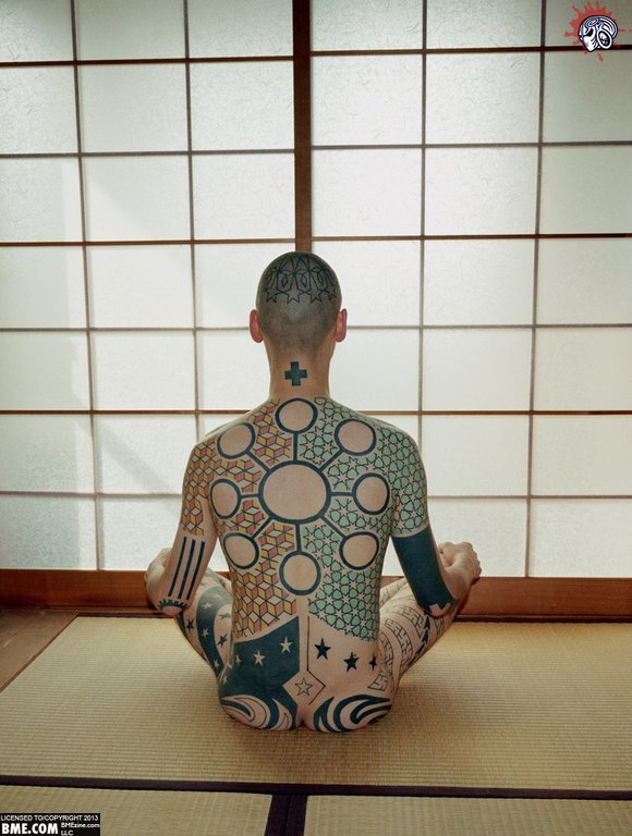 A person sits in a meditation pose, nude, facing away from the camera in a Japanese style tatami room. Their back is extensively tattooed in a symmetrical pattern of circles and contrasting colors.
