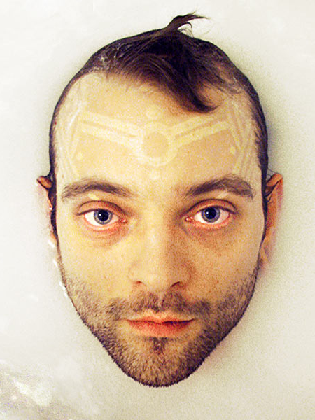 The face of Shannon Larratt, a skinny male of European descent, with brown hair, stubble and a white ink forehead tattoo coming out of what seems to be milk.
